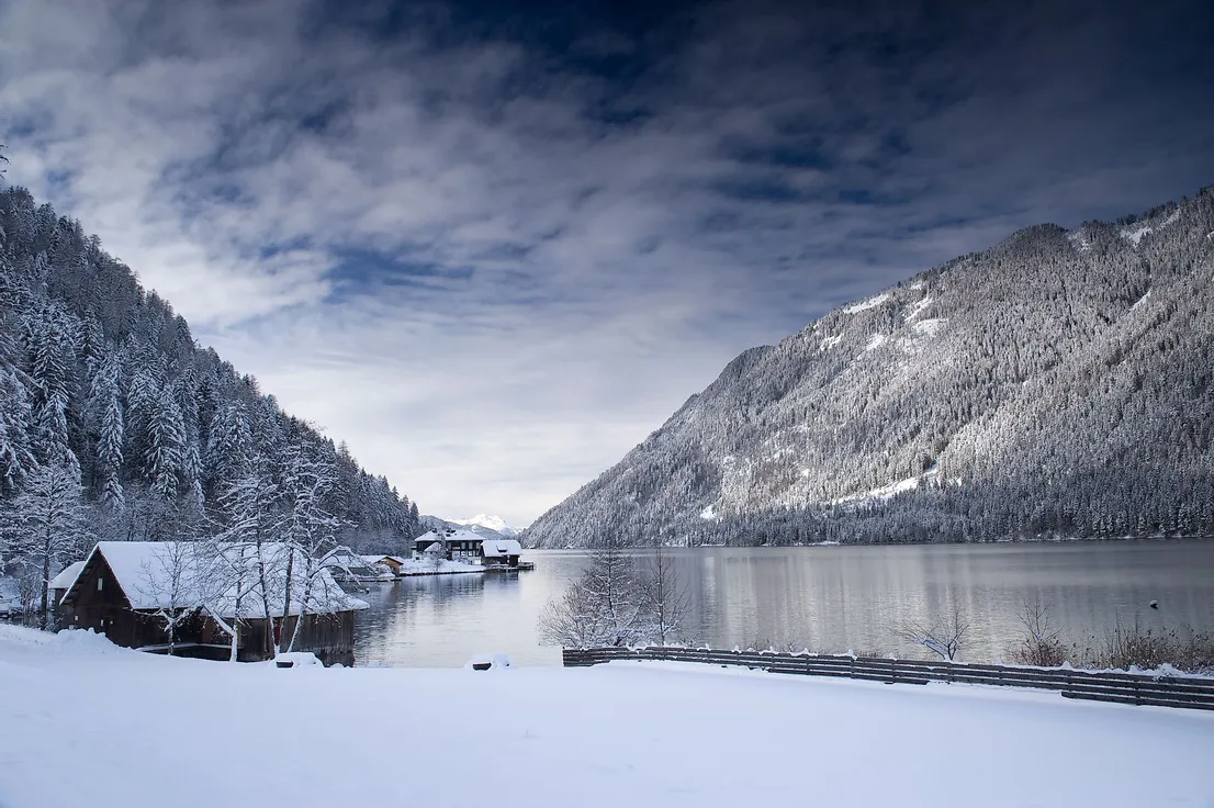 Verschneiter Alpensee mit Holzhütten und bewaldeten Bergen im Winter